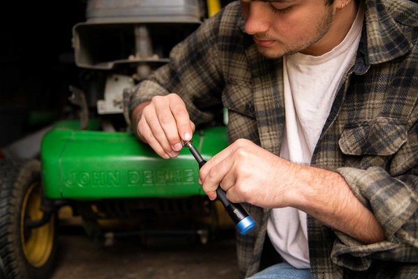 Leaning on a Deere lawnmower, a man attaches a socket to a screwdriver